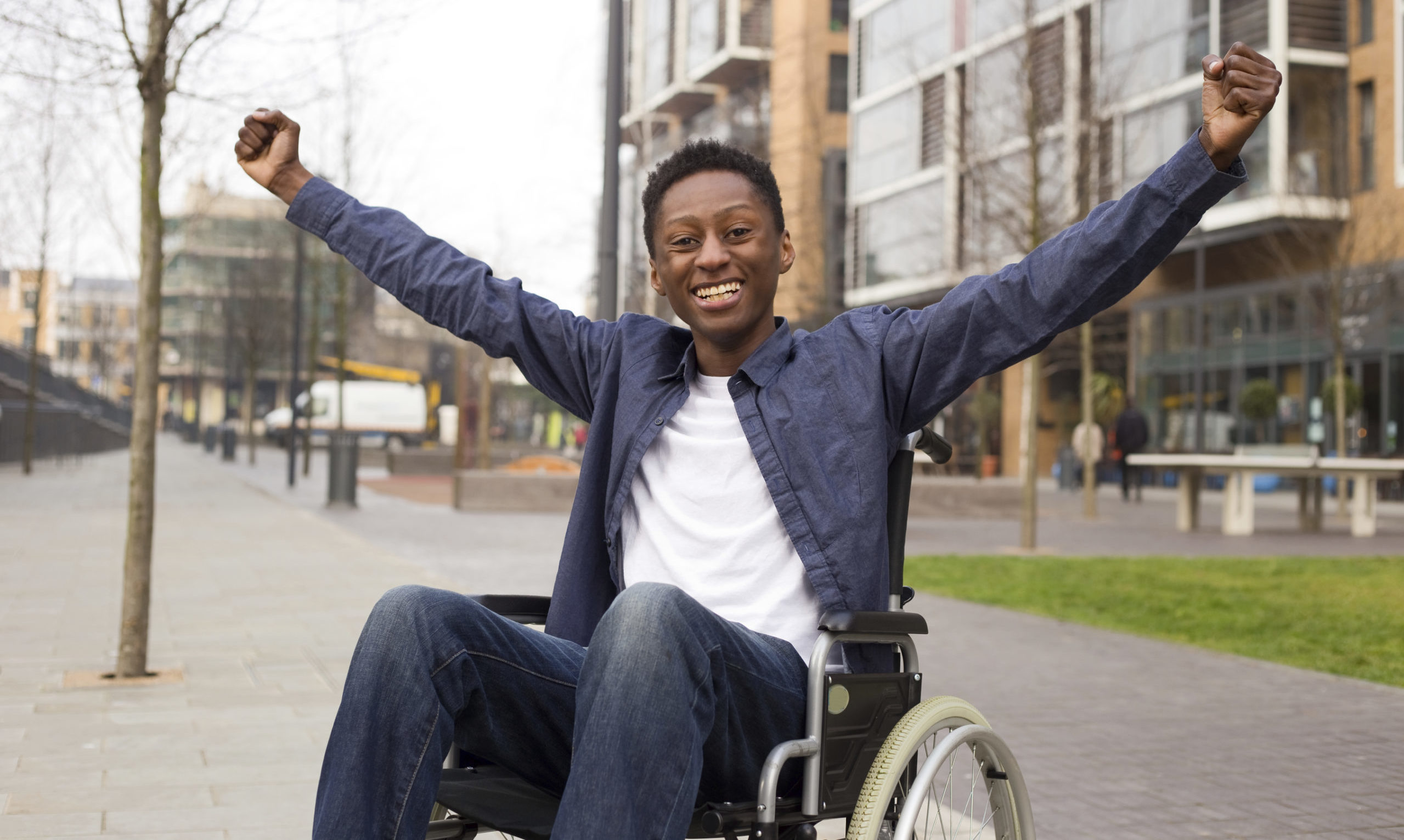 A black man smiling and wearing a white shirt and blue collar short and blue jeans in a wheelchair with his arms up and hands in a fist